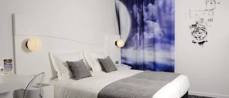 A modern hotel room with a neatly made bed, a desk with a chair, and a large wall tapestry depicting a space theme.
