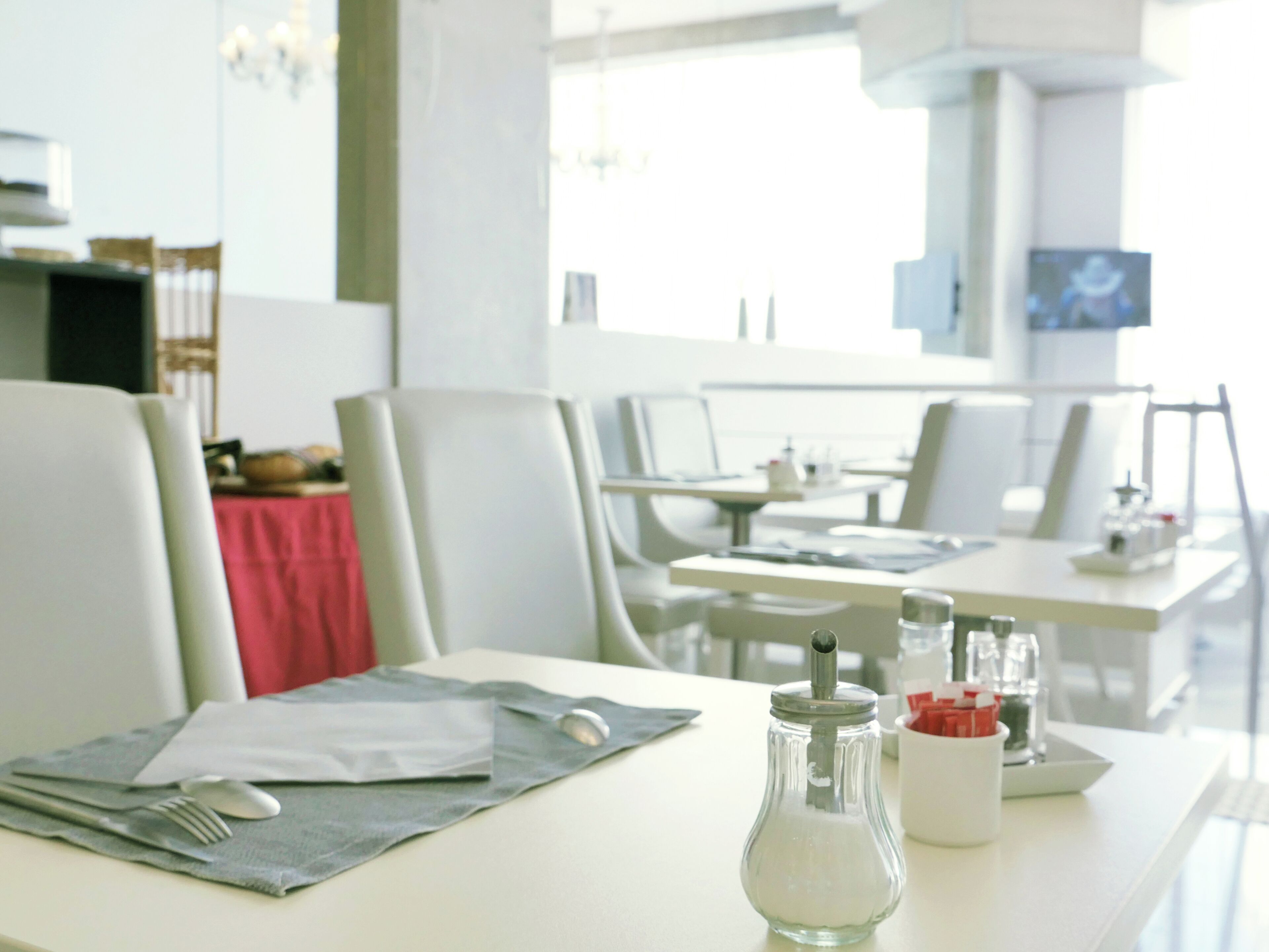 Un moderno comedor con mesas y sillas blancas, un mantel rojo y un juego de salero y pimentero sobre una mesa.