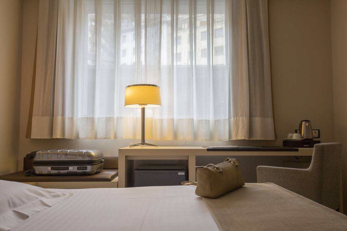 Une chambre d’hôtel avec un lit, un bureau, une lampe, une valise, un sac à main et une fenêtre avec des rideaux légers.