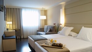 A hotel room with a large bed, a desk, a chair, and a small seating area.