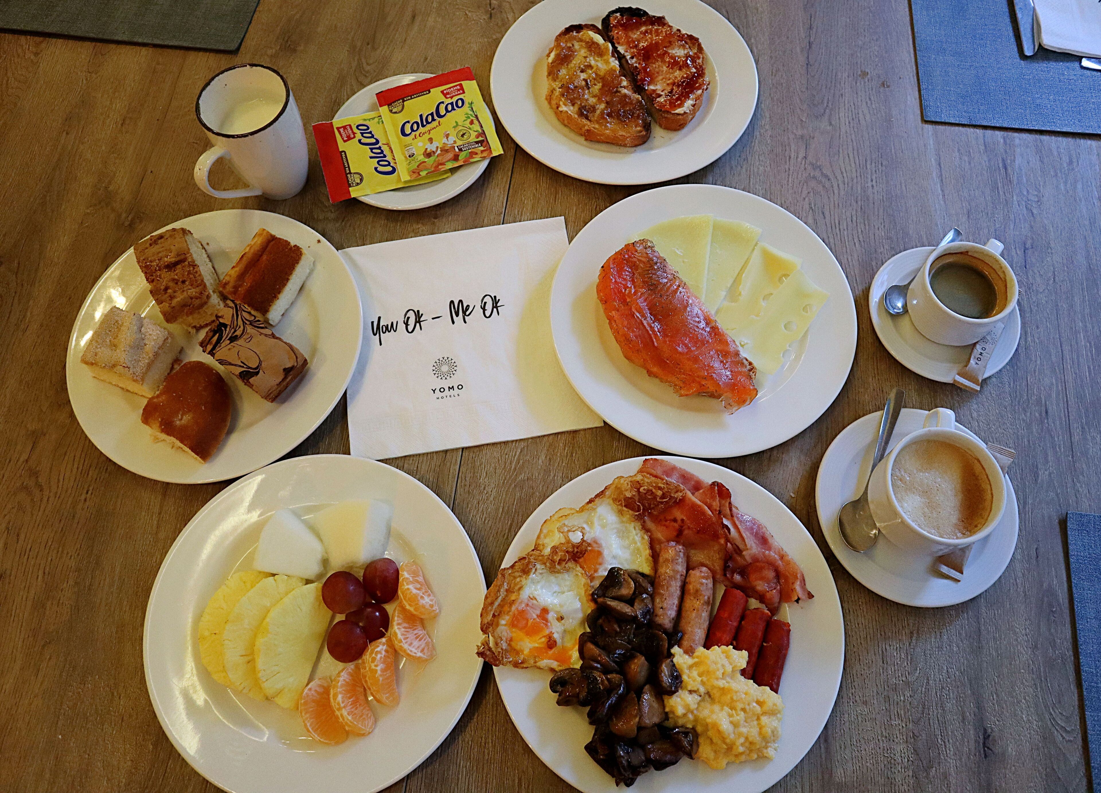 A breakfast spread on a wooden table with plates of eggs, bacon, sausages, ham, mushrooms, pineapple, and oranges; a plate of toast; a packet of Colcaao; a cup of coffee; and a napkin with "You OK - Me OK" written on it.