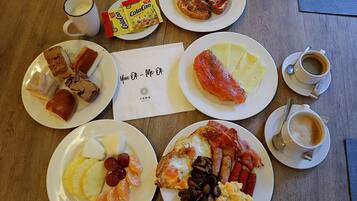 A breakfast spread on a wooden table with plates of eggs, bacon, sausages, ham, mushrooms, pineapple, and oranges; a plate of toast; a packet of Colcaao; a cup of coffee; and a napkin with "You OK - Me OK" written on it.