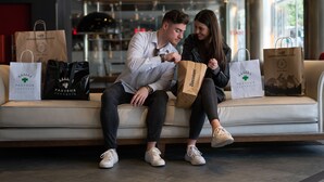 A couple sitting on a sofa with shopping bags, in a modern indoor setting with a glass partition and a visible emergency exit sign.