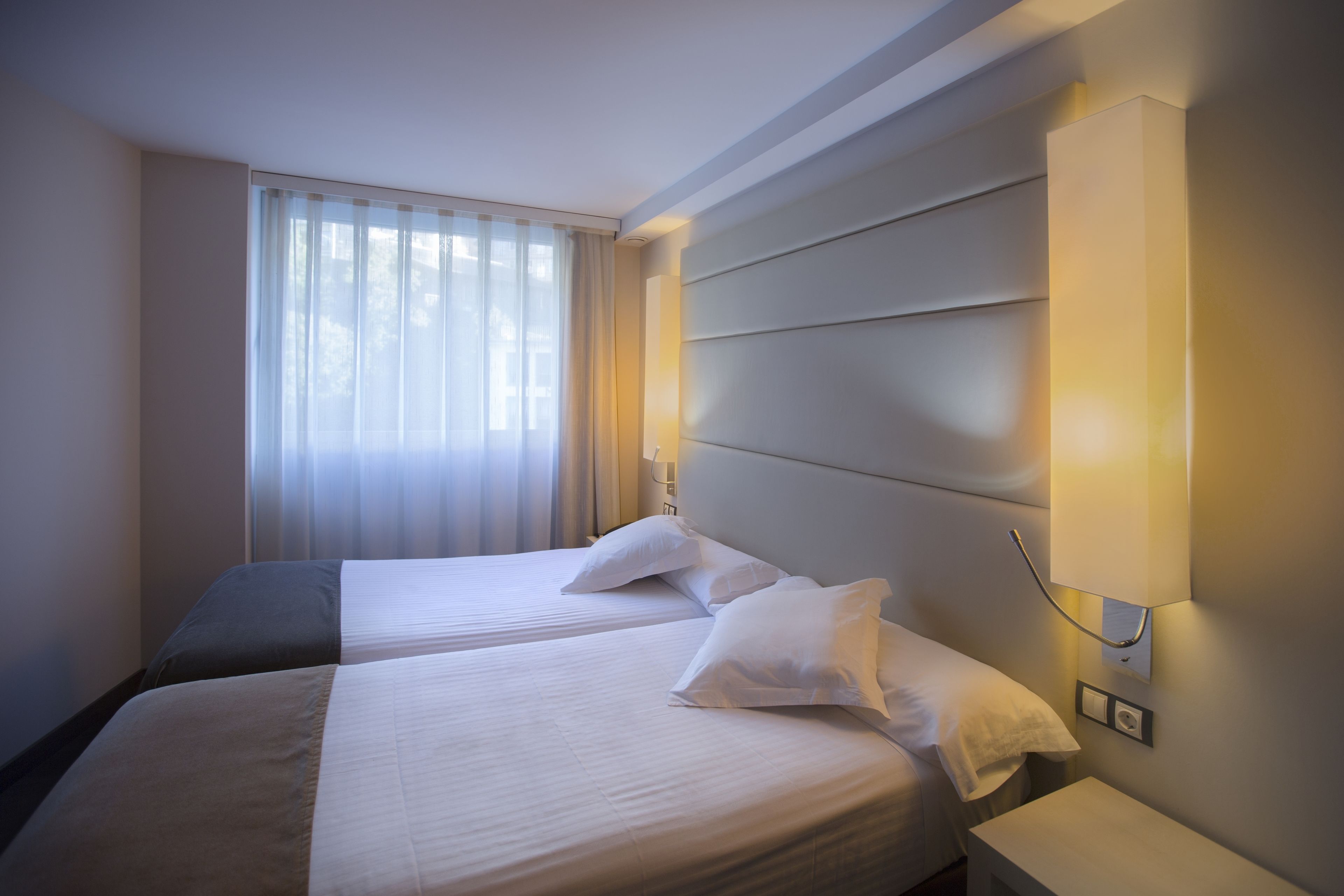 A hotel room with two beds, a bedside table, a lamp, and a window with curtains.