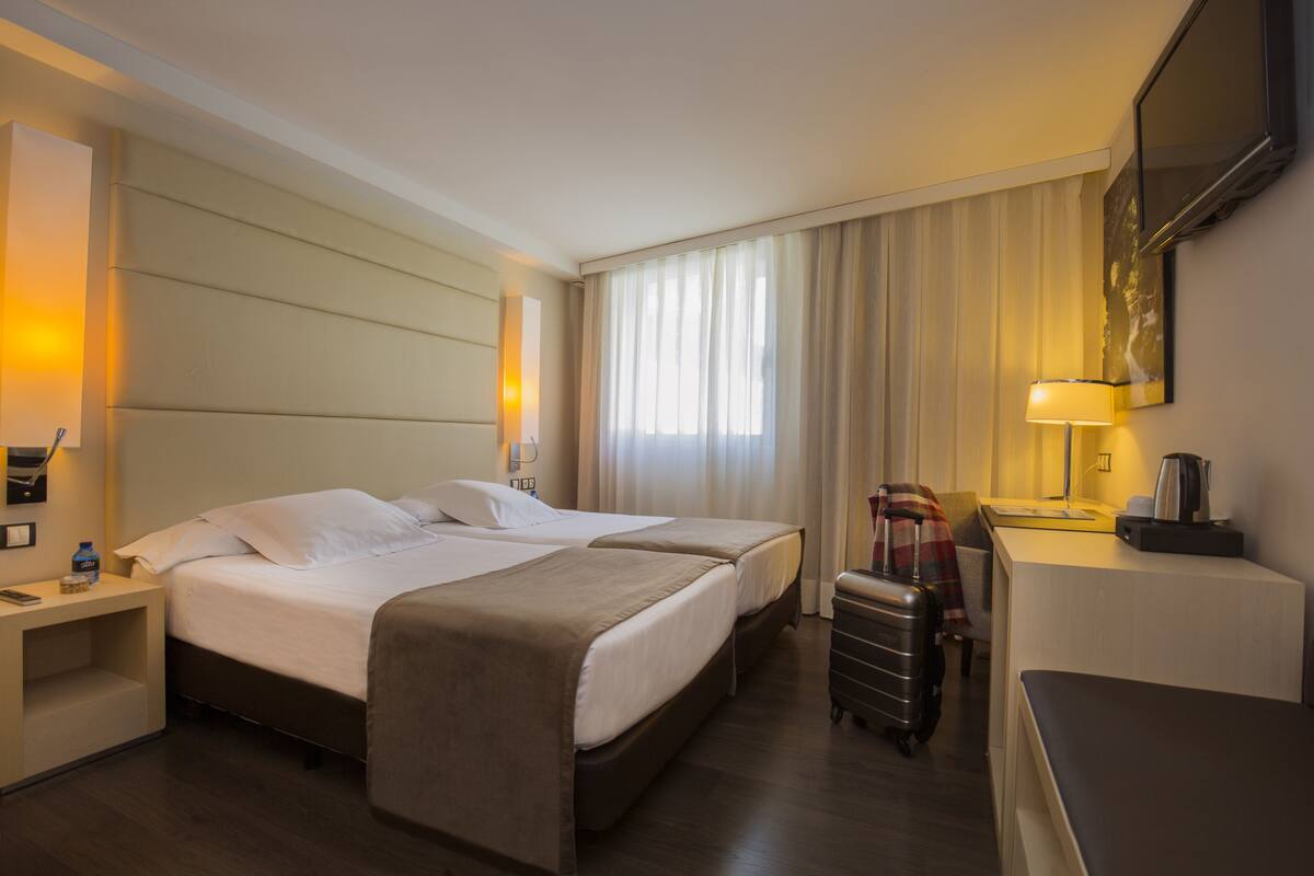 Une chambre d’hôtel avec deux lits, un bureau, une chaise, une valise et un téléviseur fixé au mur.