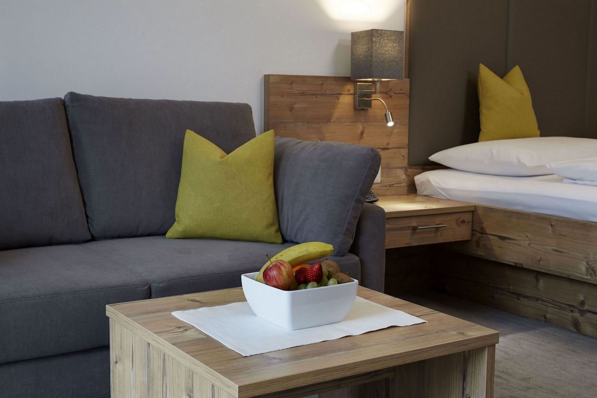Ein modernes Hotelzimmer mit grauem Sofa, einem Holfnachtkästchen mit Lampe und einem Bett mit weißer Bettwäsche.