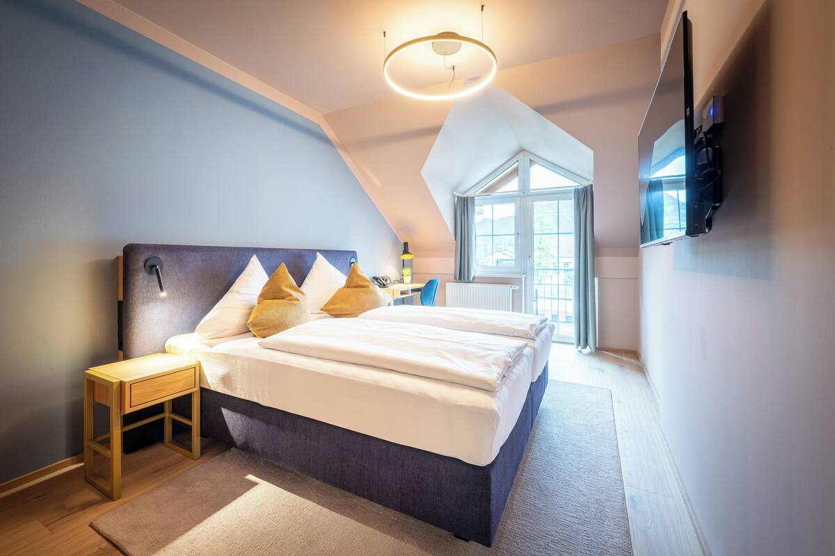 Ein modernes Schlafzimmer mit einem großen Bett, einem Nachttisch, einem an der Wand montierten Fernseher und einem Fenster, das natürliches Licht hereinlässt.