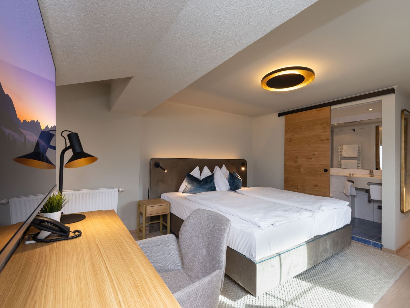 A modern bedroom with a bed, bedside table, lamp, and a view of a mountain at sunset.