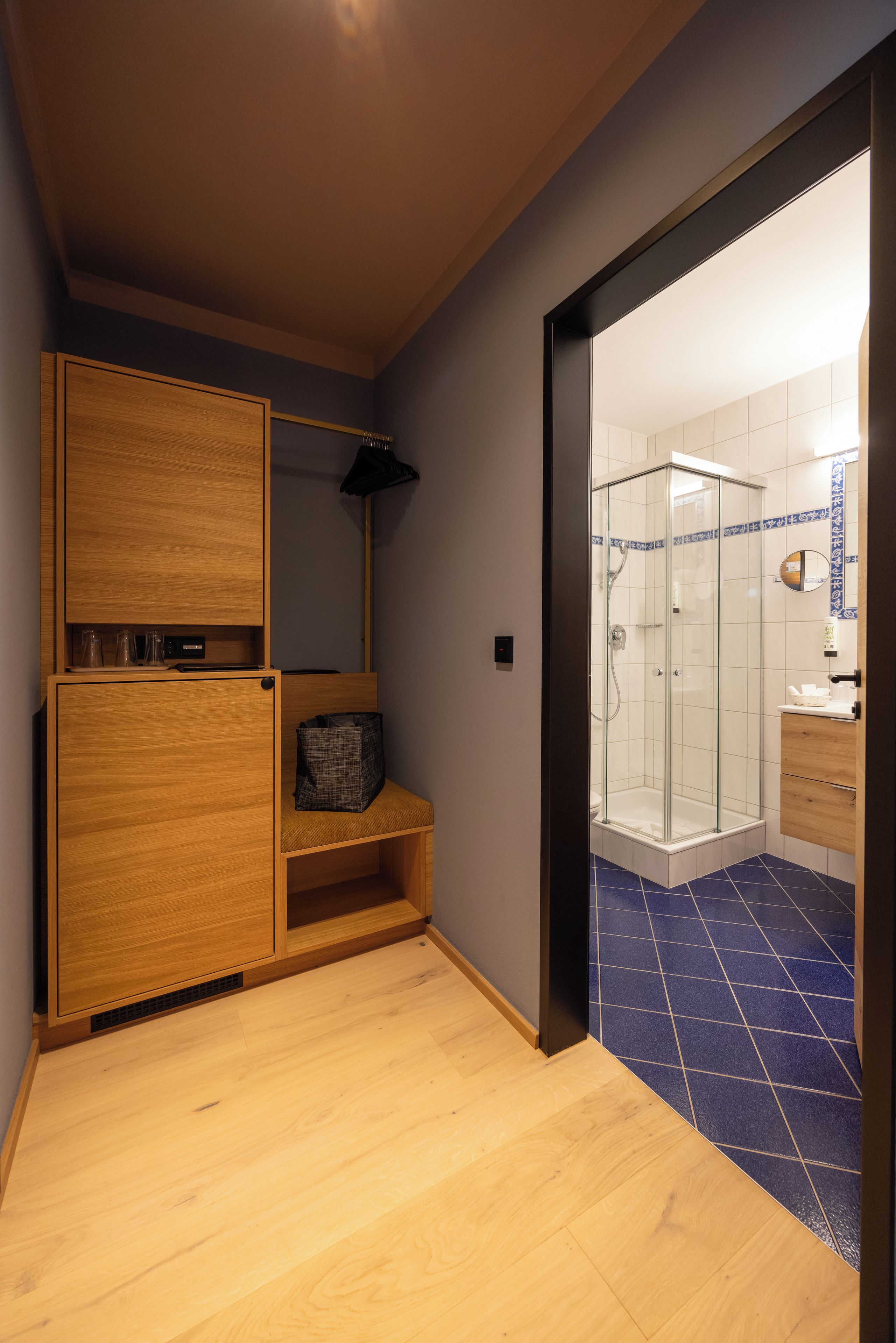 Une chambre d’hôtel avec une armoire en bois, un banc, une petite table et une cabine de douche en verre.