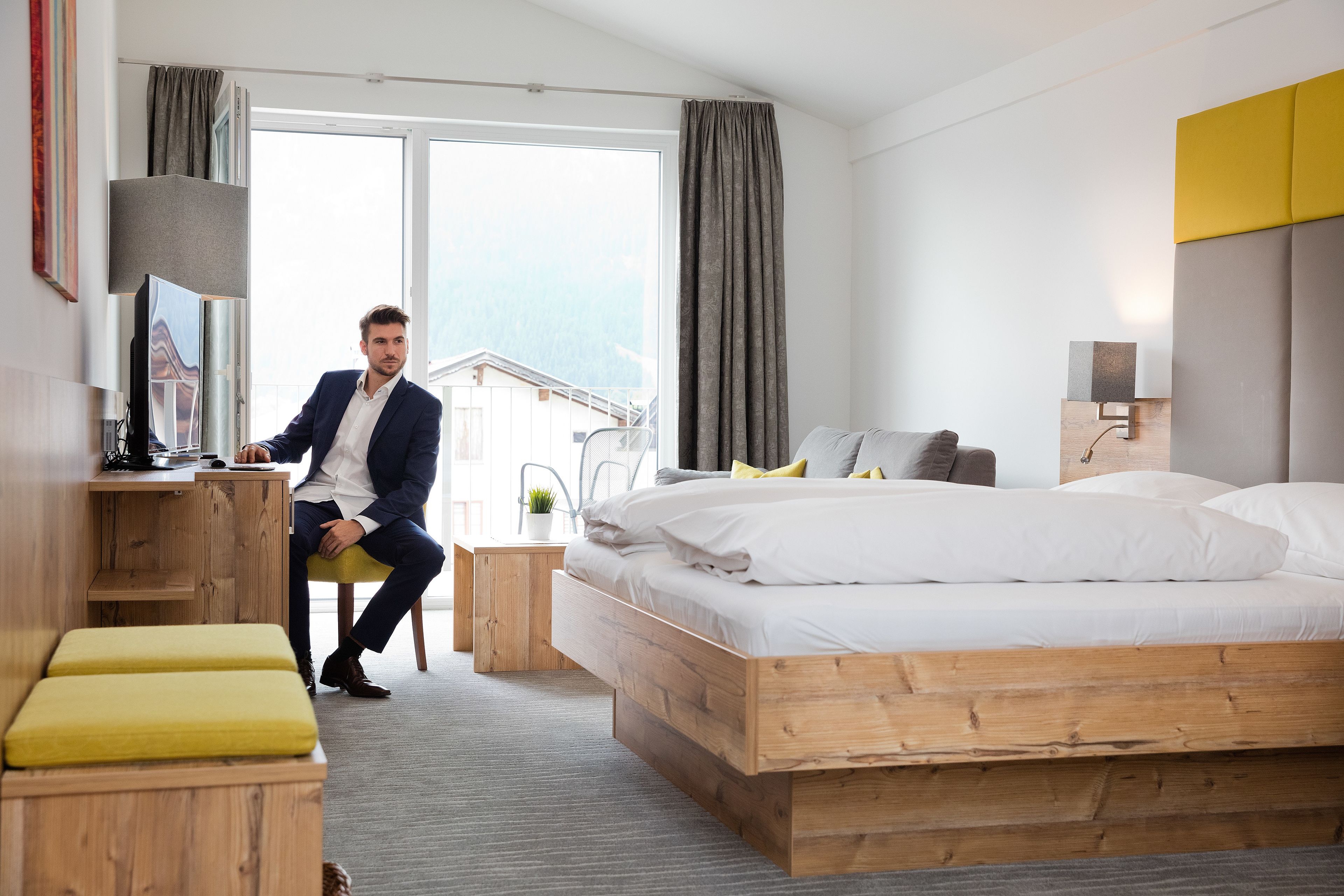 A modern hotel room with a wooden bed, a desk, a chair, and a view of the mountains.