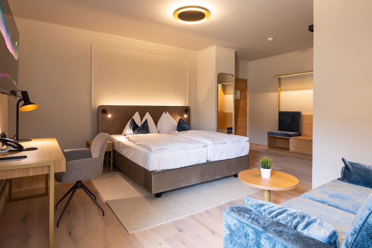 Ein modernes Hotelzimmer mit einem großen Bett, einem Schreibtisch mit Lampe, einem kleinen runden Tisch mit Pflanze und einer Sitzecke mit Sofa.