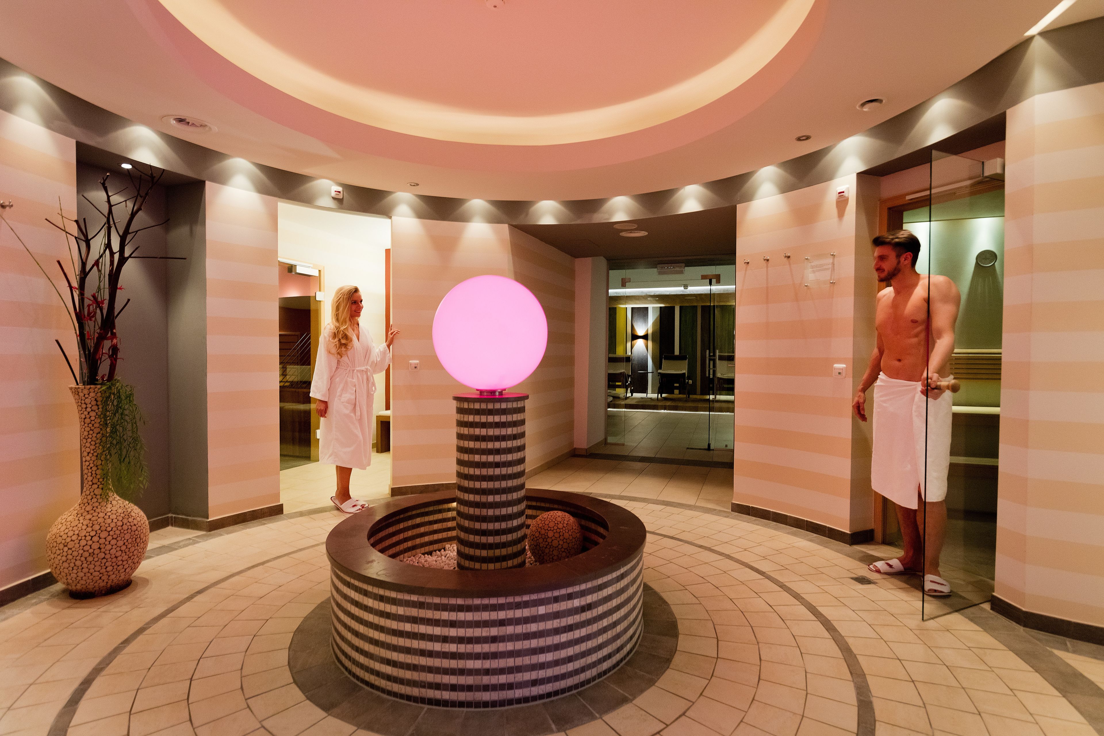 Un lobby moderno con una zona de estar circular central, un jarrón grande con ramas y una lámpara esférica rosa.