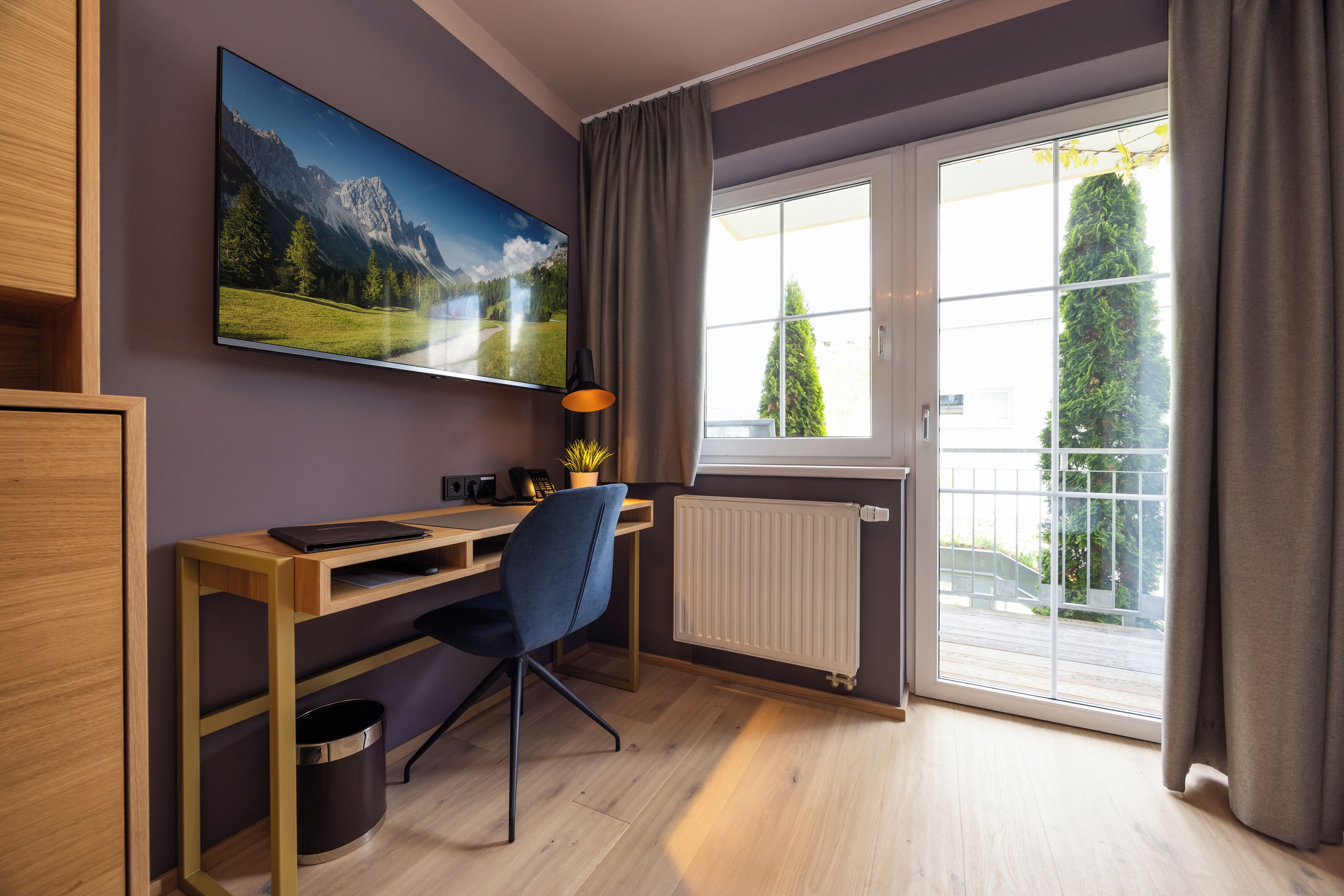 A modern room with a wooden desk, a chair, a mounted TV, a radiator, and a window with curtains.