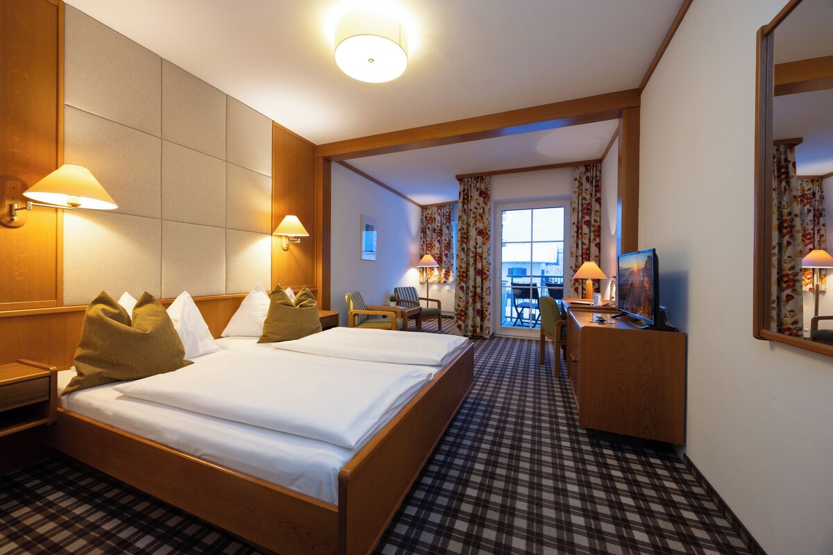 A hotel room with a large bed, two bedside lamps, a desk with a chair, a television, and a view of the outside through a window.