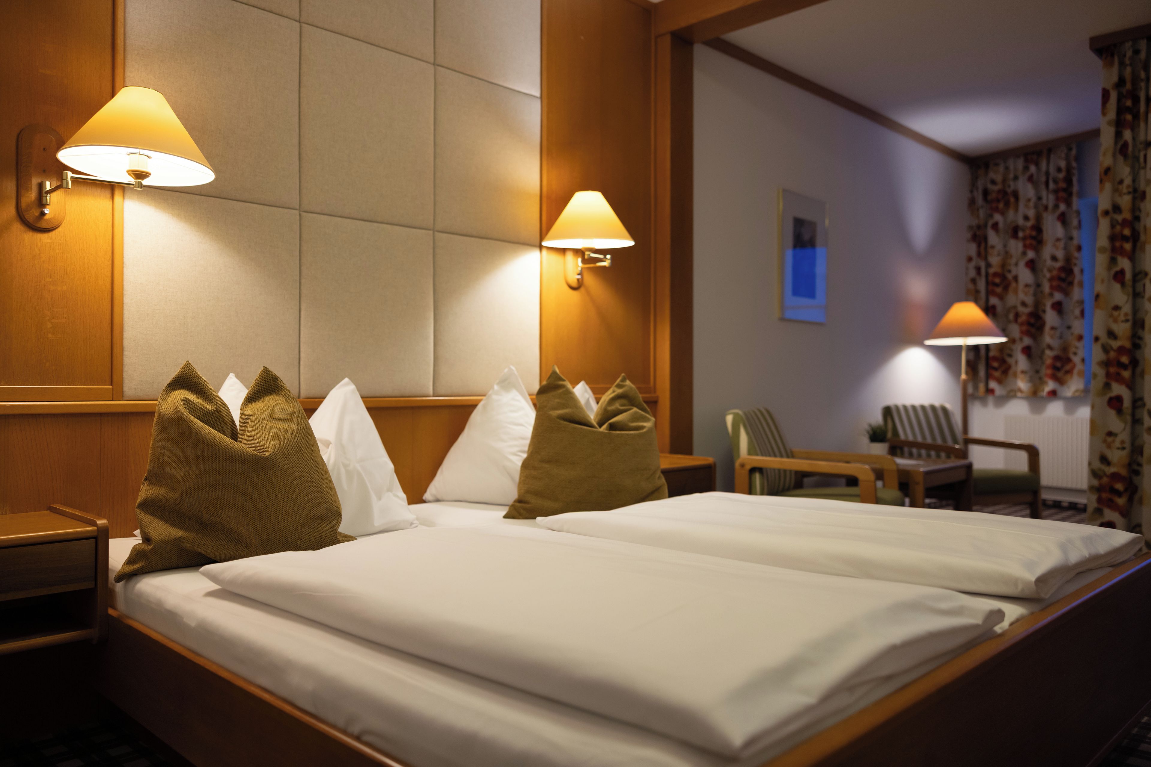 A hotel room with a neatly made bed, two wall-mounted lamps, a wooden nightstand, a chair, and a window with curtains.