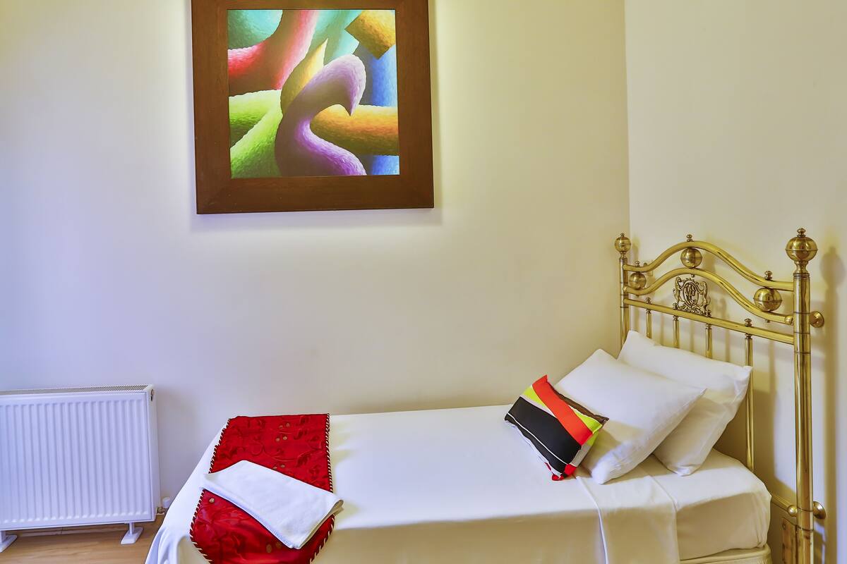 A single bed with a gold headboard, white bedding, and a colorful pillow. A radiator is on the floor beside the bed.