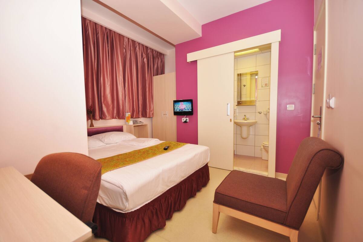 A hotel room with a bed, a desk, a chair, a TV, and a bathroom visible through an open door.