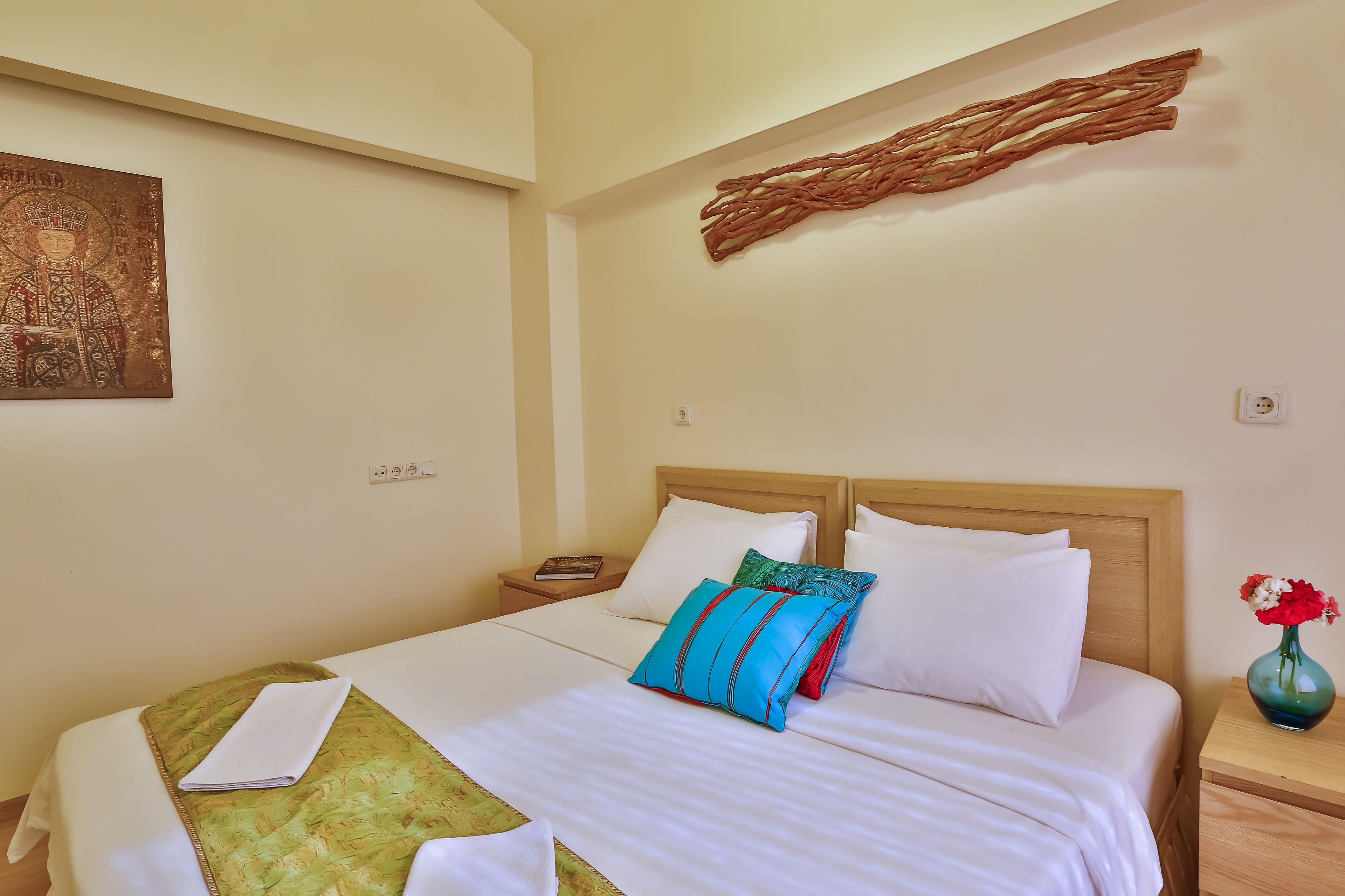 A neatly made bed with white linens and multiple pillows, a bedside table with a book and a vase of flowers, and a wall-mounted artwork.