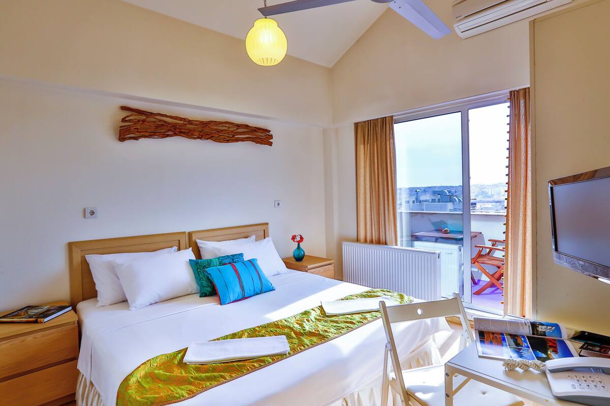 A hotel room with a bed, bedside table, chair, and a view of the city through a large window.