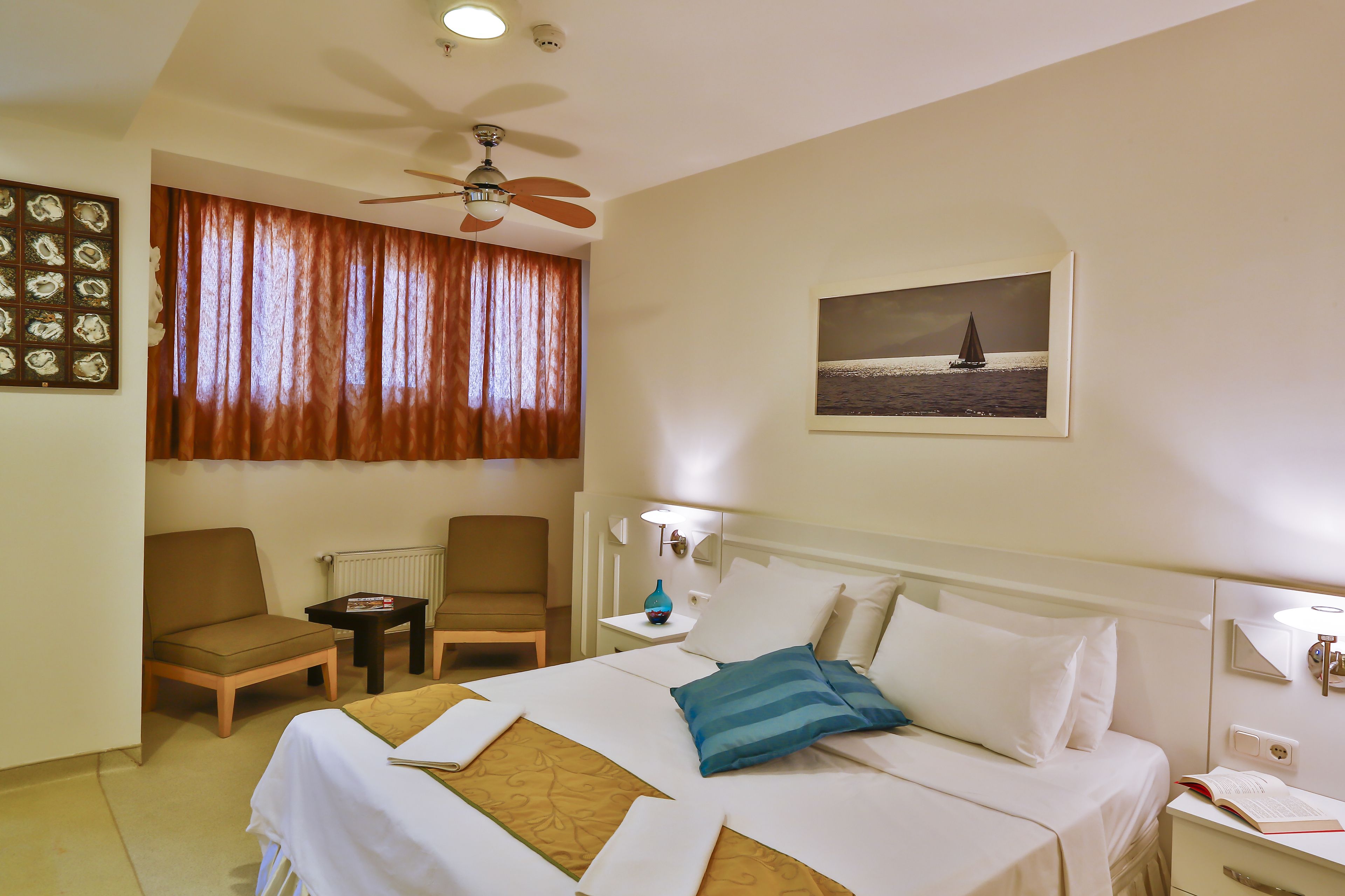 A hotel room with a bed, two chairs, a small table, a ceiling fan, a painting, and a nightstand with a lamp.