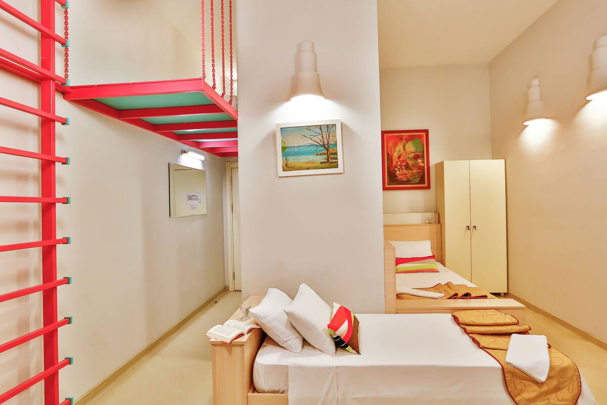 A hotel room with a bed, a small desk with a chair, a wardrobe, and a red staircase.