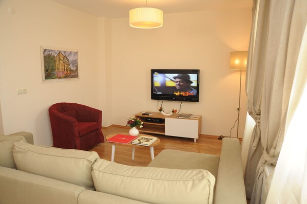 A living room with a television, a red armchair, a white sofa, a wooden coffee table with books and a vase, and a painting on the wall.