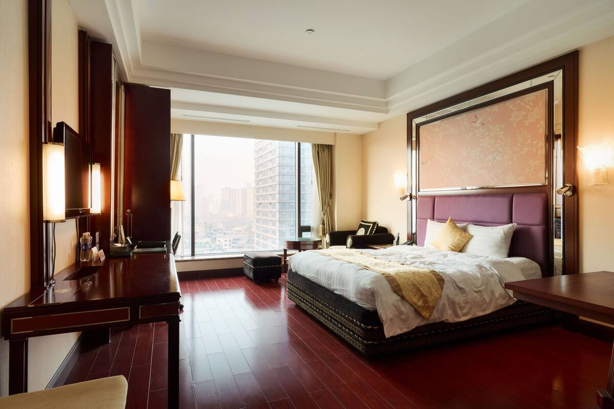 Een hotelkamer met een groot bed, een bureau, een stoel, een nachtkastje en een groot raam met uitzicht over de stad.