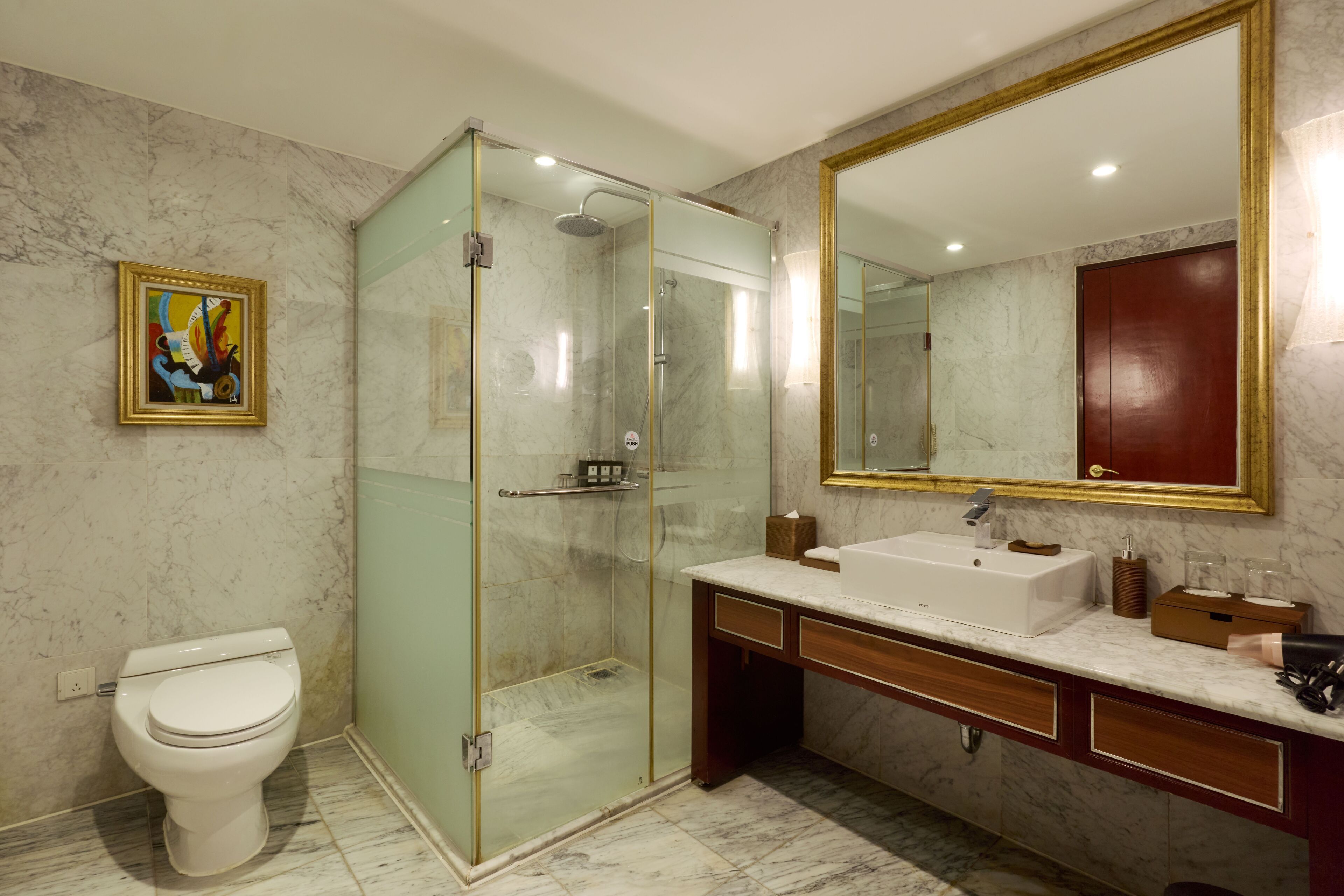 Et moderne badeværelse med bruseniche, et hvidt toilet, et marmorgulv, et stort spejl med gylden ramme og et træbord med vask.