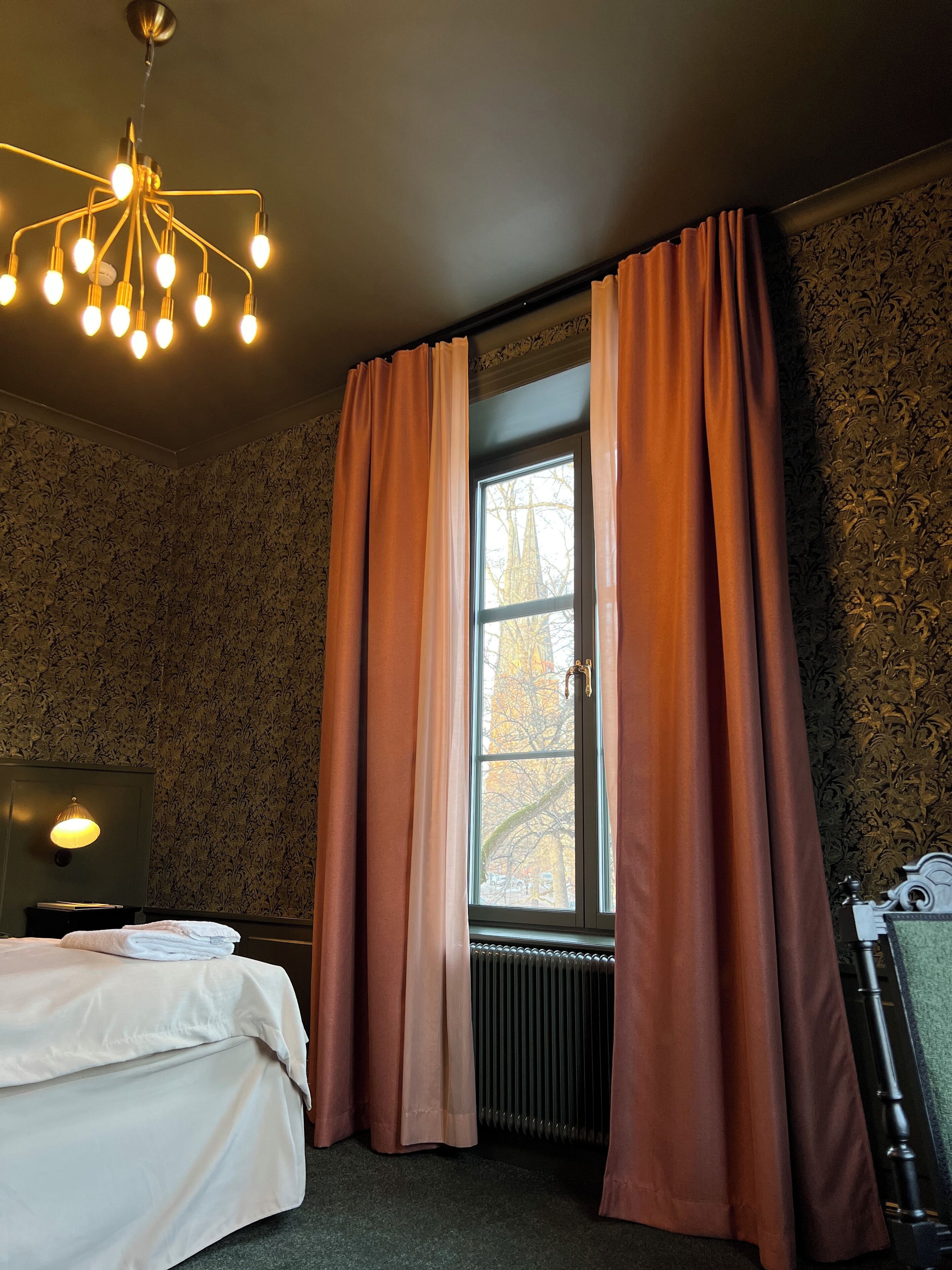 A hotel room with a bed, a radiator, a window with curtains, a wall with a floral pattern, and a chandelier.