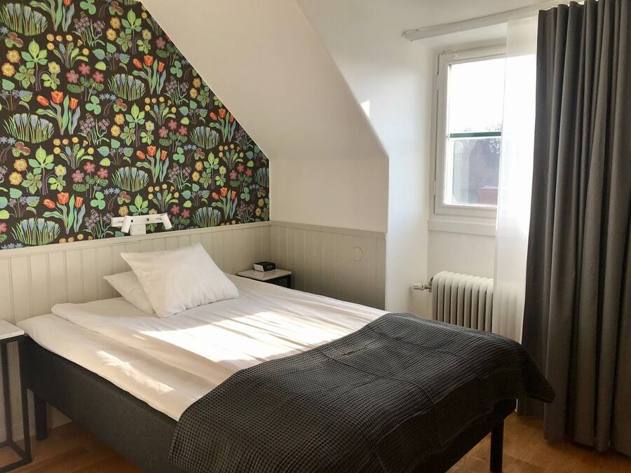 Een slaapkamer met een bed, nachtkastje, een wandklok, een radiator en een raam met gordijnen.