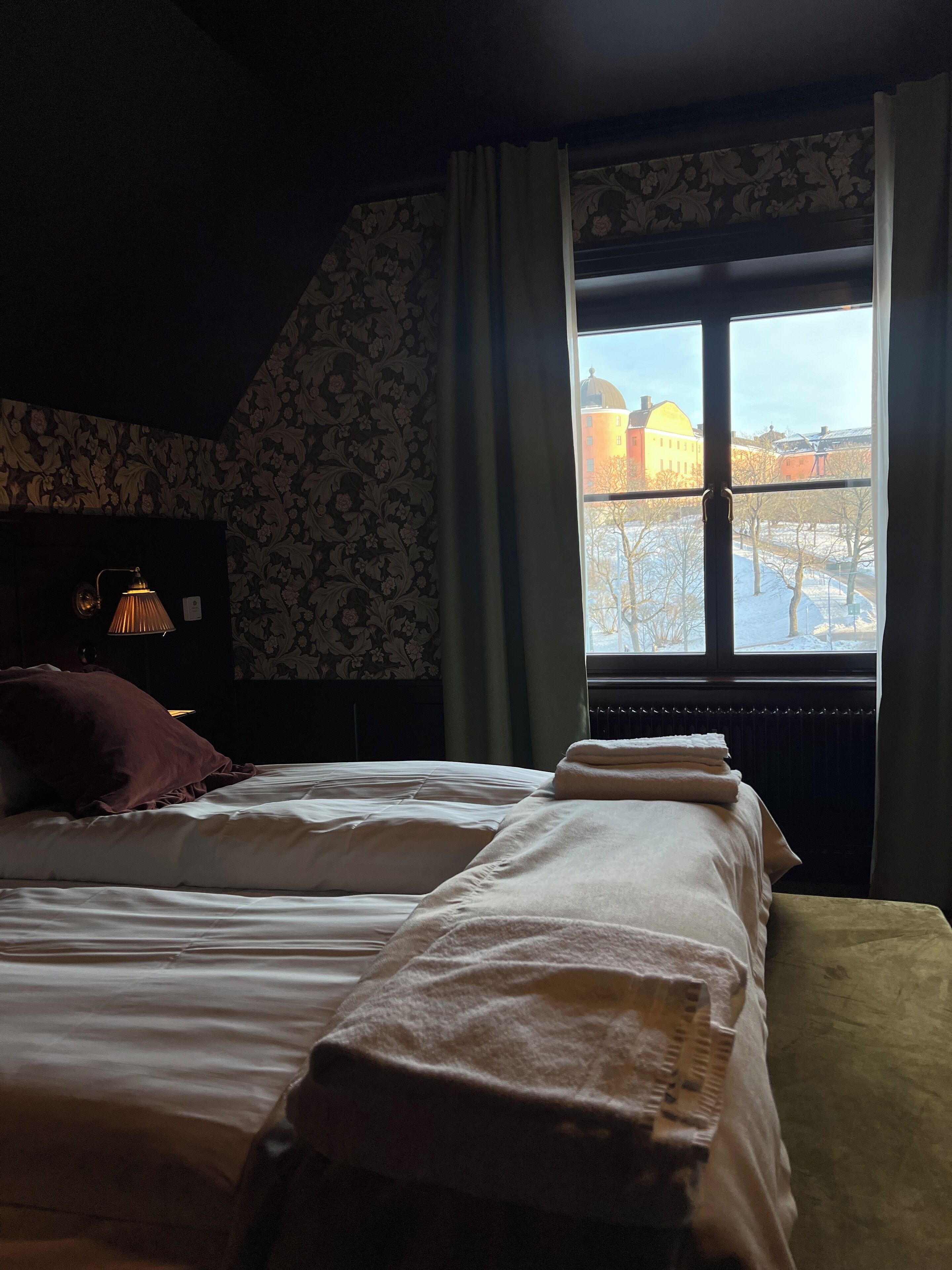 Un dormitorio con dos camas, un radiador, una lámpara y una ventana con vistas a edificios.