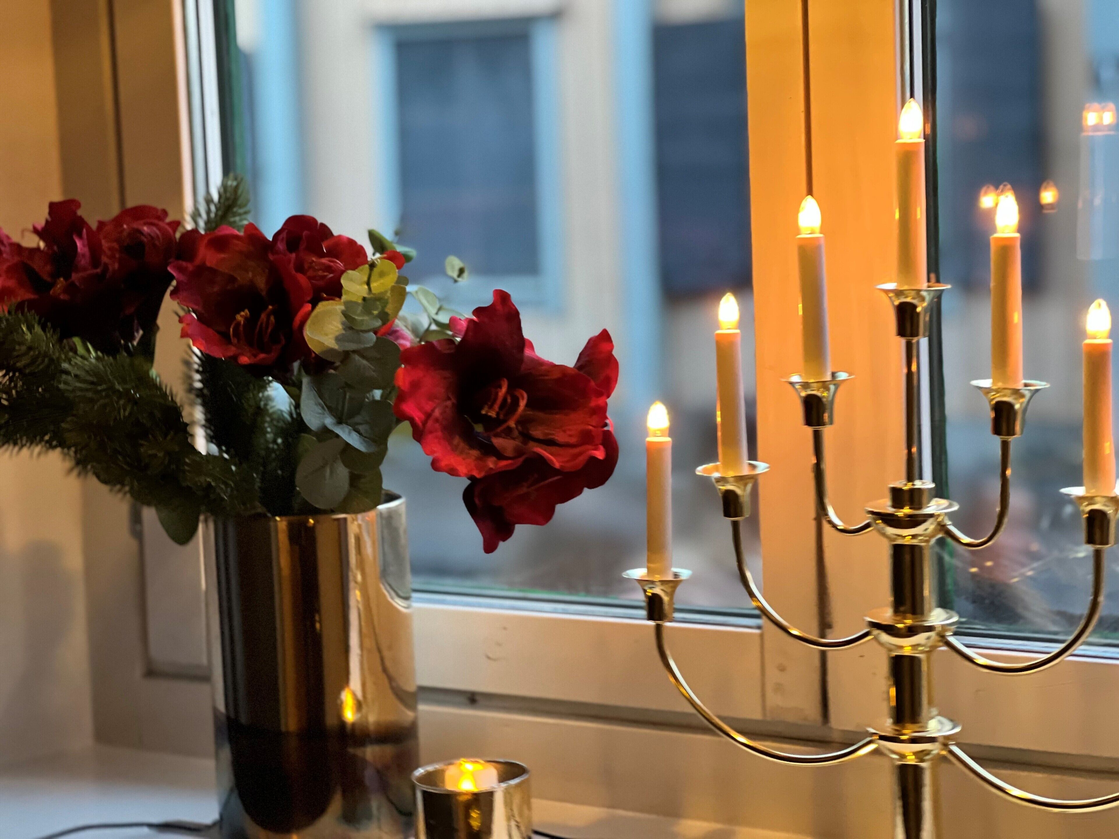 Un candelabro con velas encendidas y un florero con flores rojas sobre un alféizar.