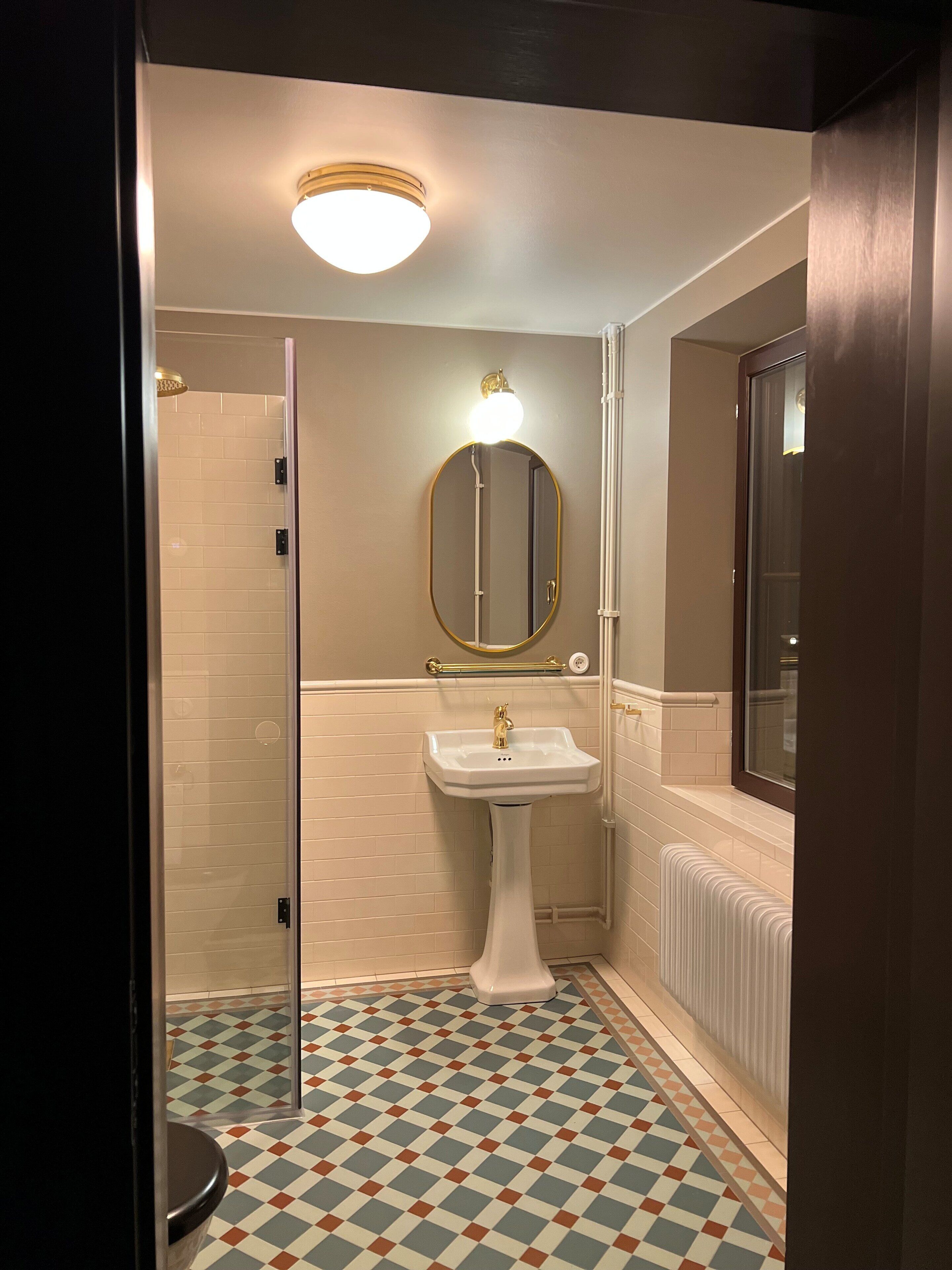 Um banheiro com piso ladrilhado, pia de pedestal, espelho com moldura dourada, box de chuveiro e radiador.