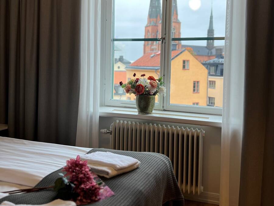 Habitación de hotel con una cama, una ventana que ofrece vistas de una iglesia y edificios, un radiador y un florero con flores en el alféizar.