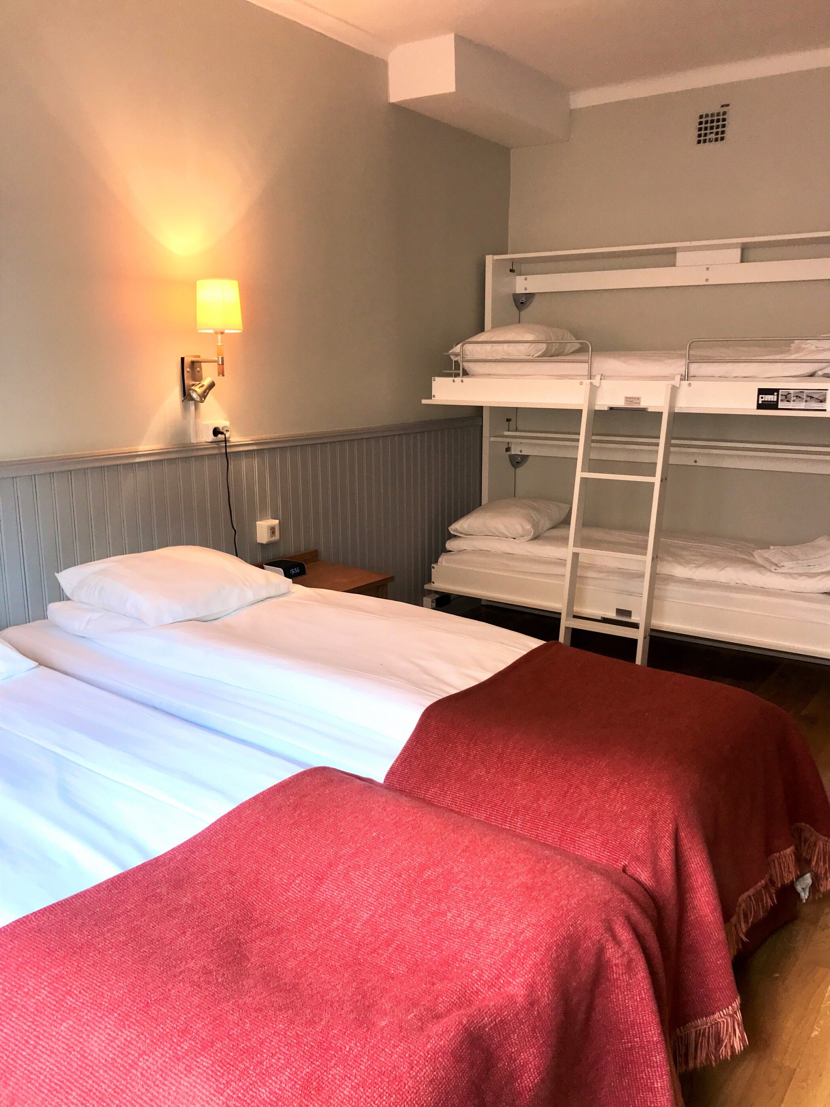 Een hotelkamer met twee bedden, waarvan één met een rode deken en de ander met witte lakens. Er is een wandlamp en een stapelbed met witte lakens.