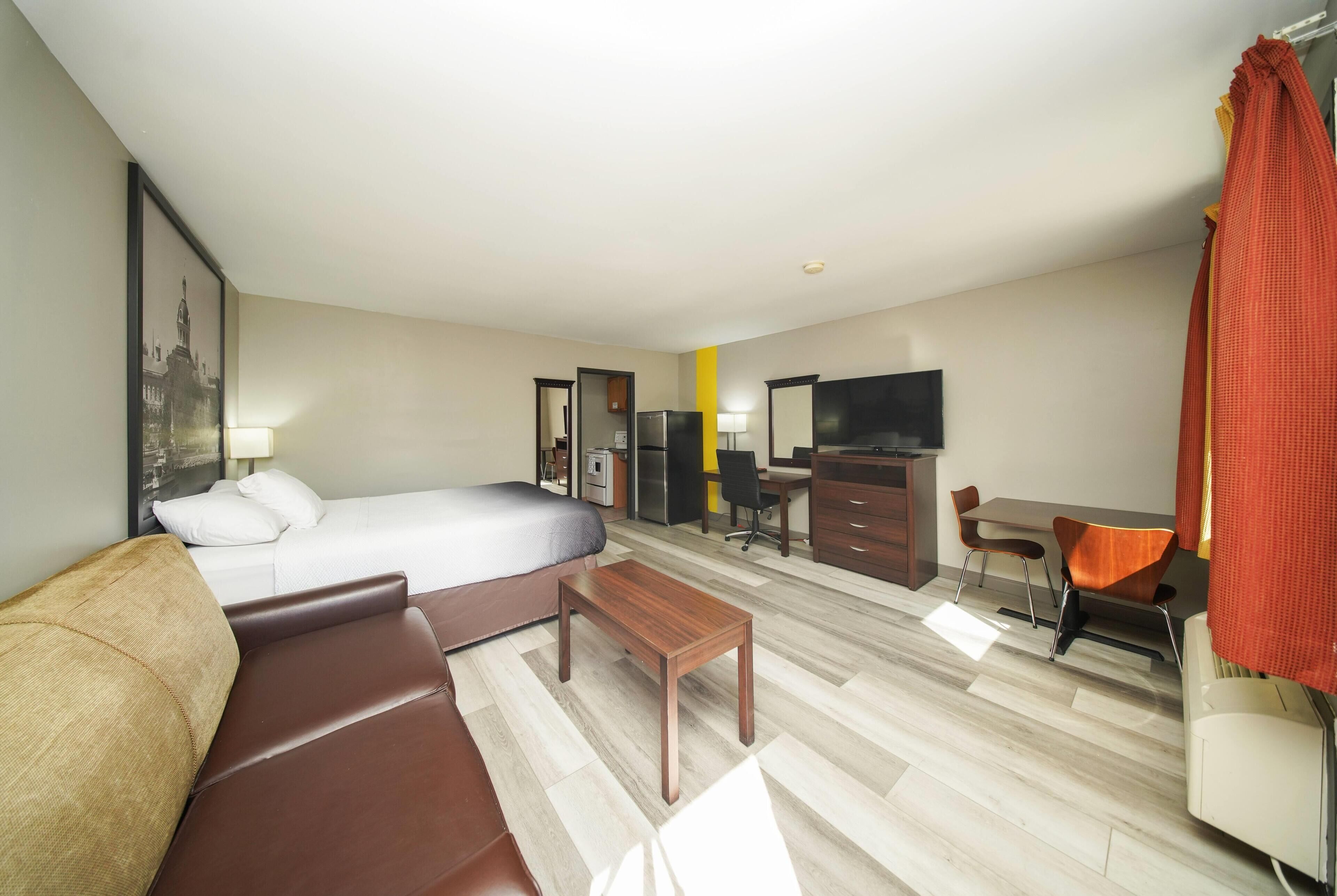 Quarto de hotel com cama, sofá, escrivaninha, cadeira, televisão e espelho.