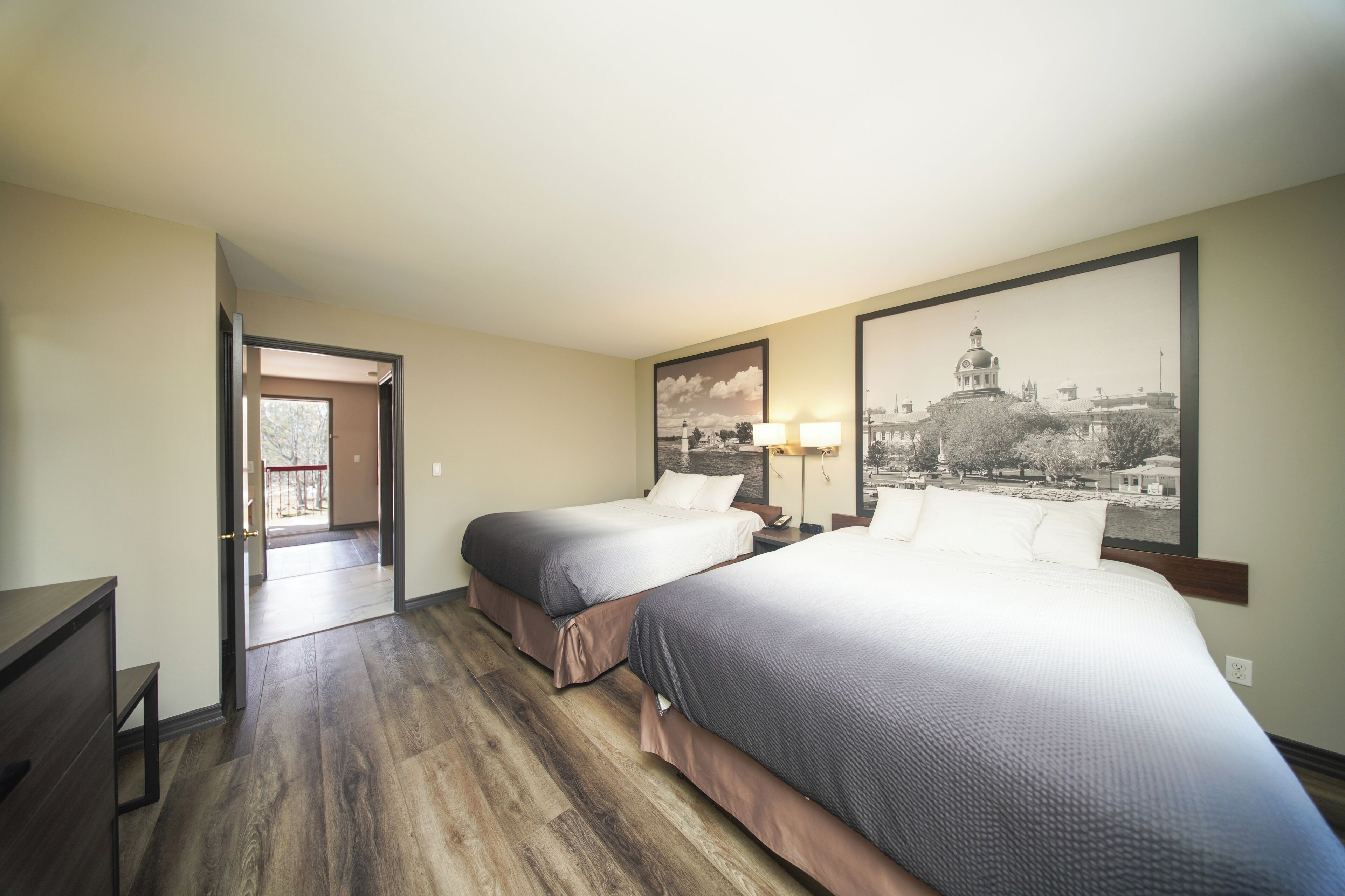 A hotel room with two beds, a desk, a chair, a door leading to a balcony, and a large framed cityscape picture on the wall.