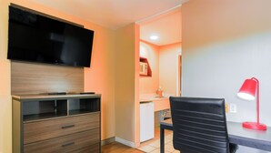 Een compacte kamer met een flatscreen televisie bevestigd aan een houten kast, een bureau met een rode bureaulamp en een zwarte stoel.