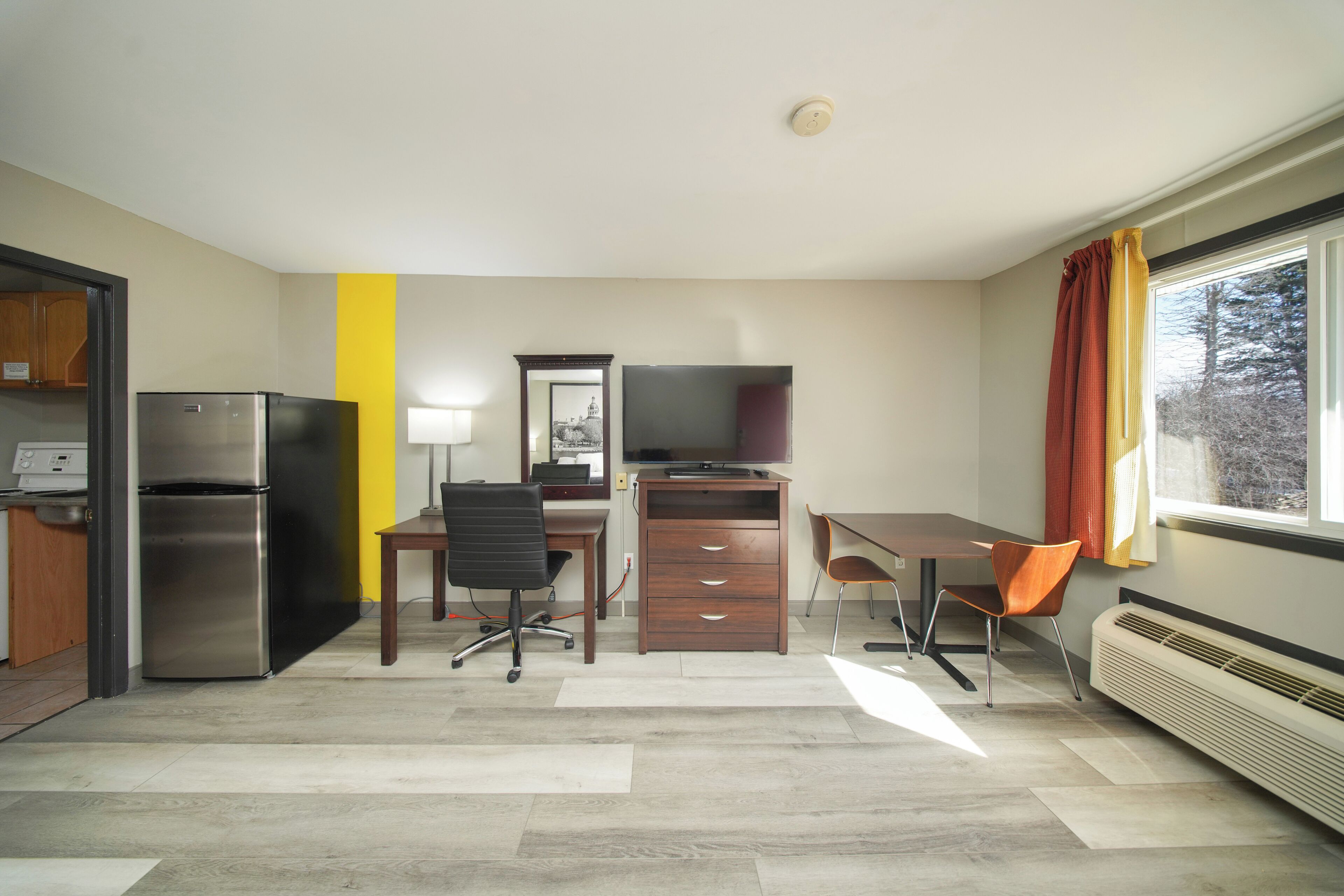 A compact room with a kitchenette, a desk with a chair, a television, and a window with curtains.