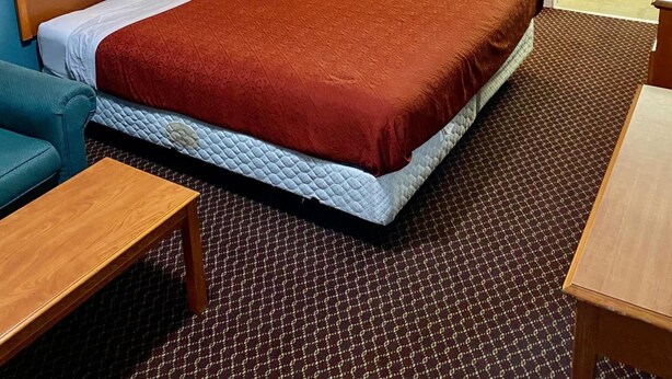 A hotel room with a bed, a nightstand with a lamp, a wooden headboard, a blue armchair, a wooden table, and a bathroom visible through an open door. - Plaza Inn Springfield (Springfield)