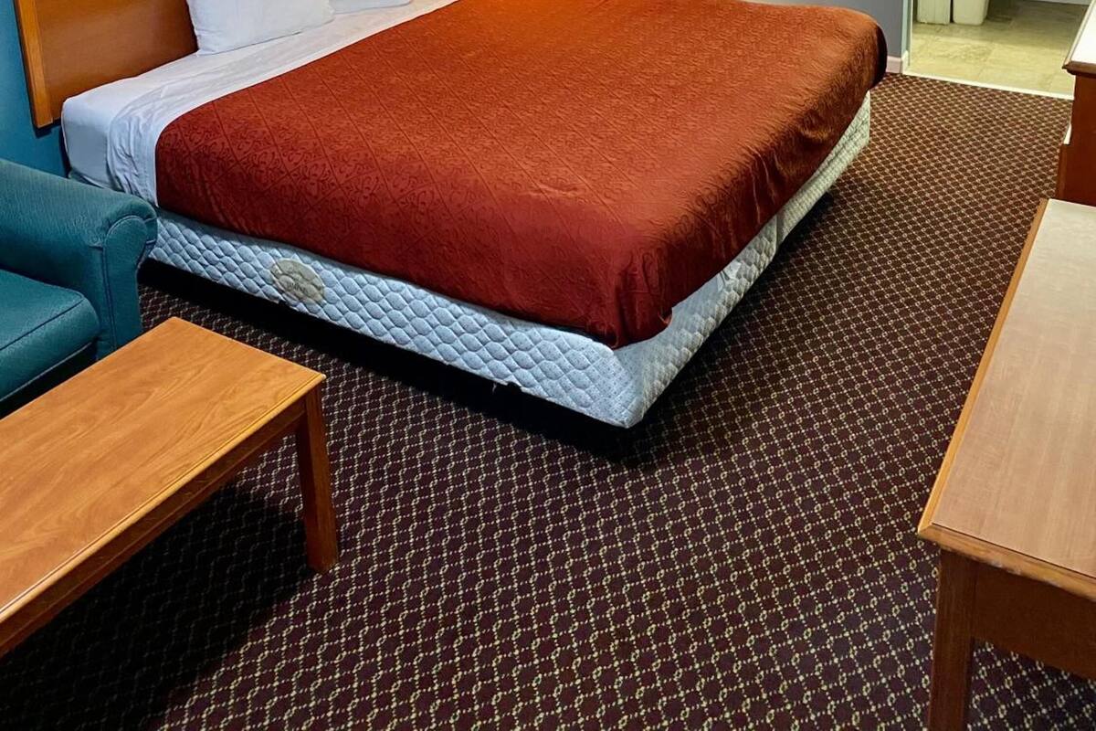 A hotel room with a bed, a nightstand with a lamp, a wooden headboard, a blue armchair, a wooden table, and a bathroom visible through an open door.
