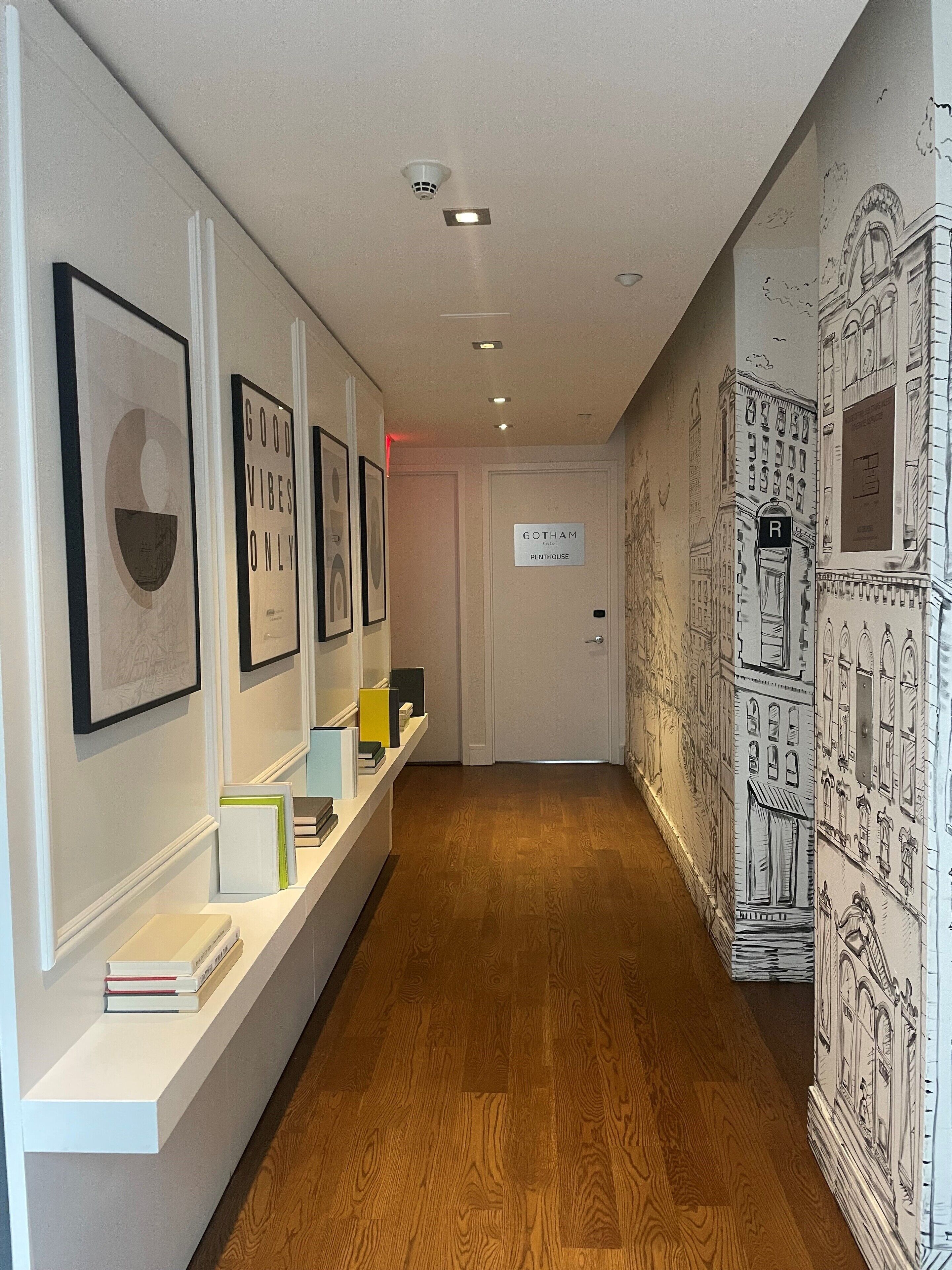 A hallway with framed artwork on the walls, a sign for 'Gotham Penthouse', and a wooden floor.
