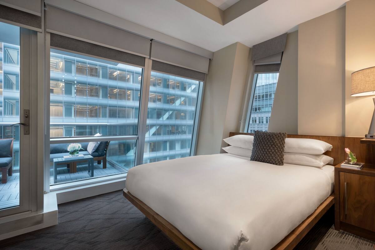 A hotel room with a large bed, a view of a building through floor-to-ceiling windows, and a small seating area with a table and chairs.