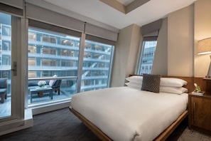 Une chambre d’hôtel avec un grand lit, une vue sur un immeuble grâce à de grandes fenêtres du sol au plafond, et un petit coin salon avec une table et des chaises.