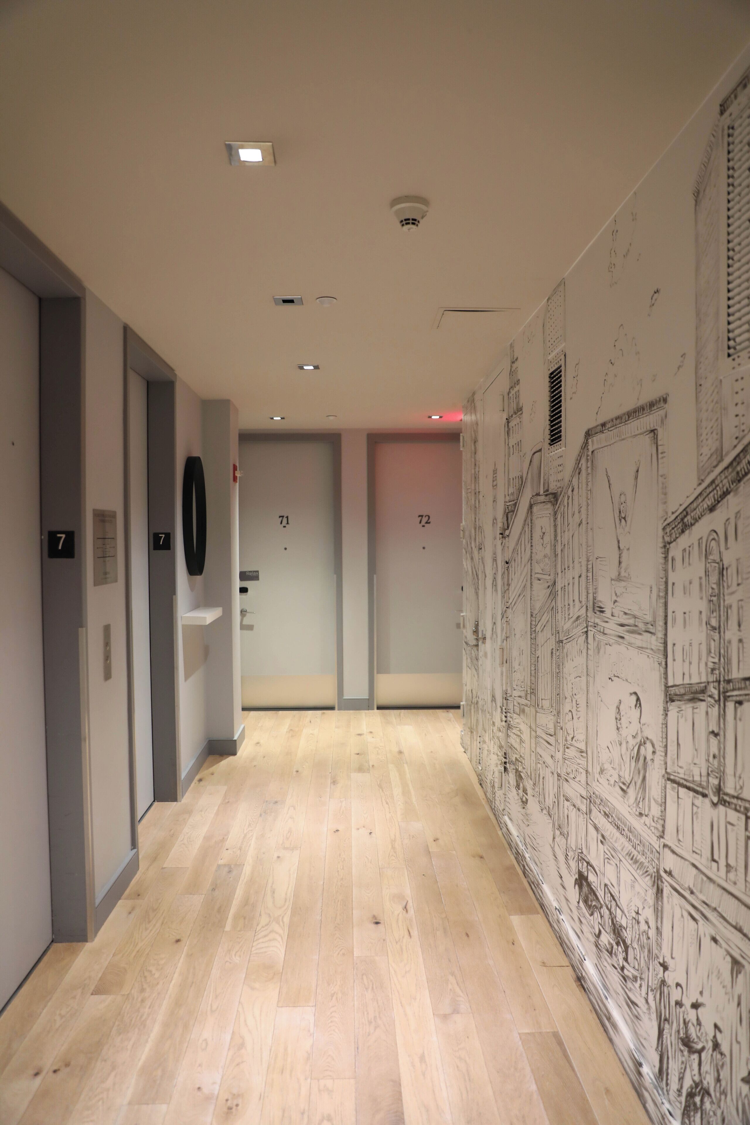 A hallway with wooden flooring, numbered doors, and a wall with architectural drawings.