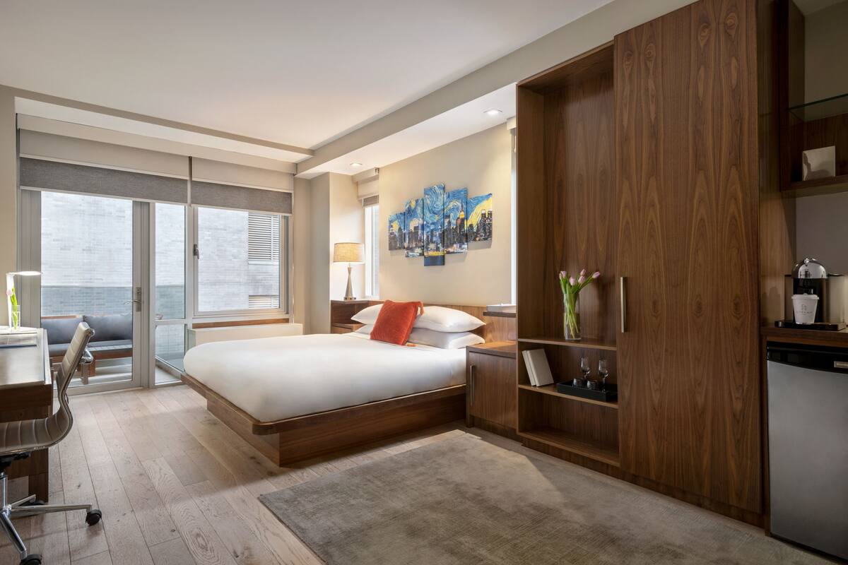 A modern hotel room with a large bed, a desk, a chair, a wardrobe, and a view of the sea through large windows.