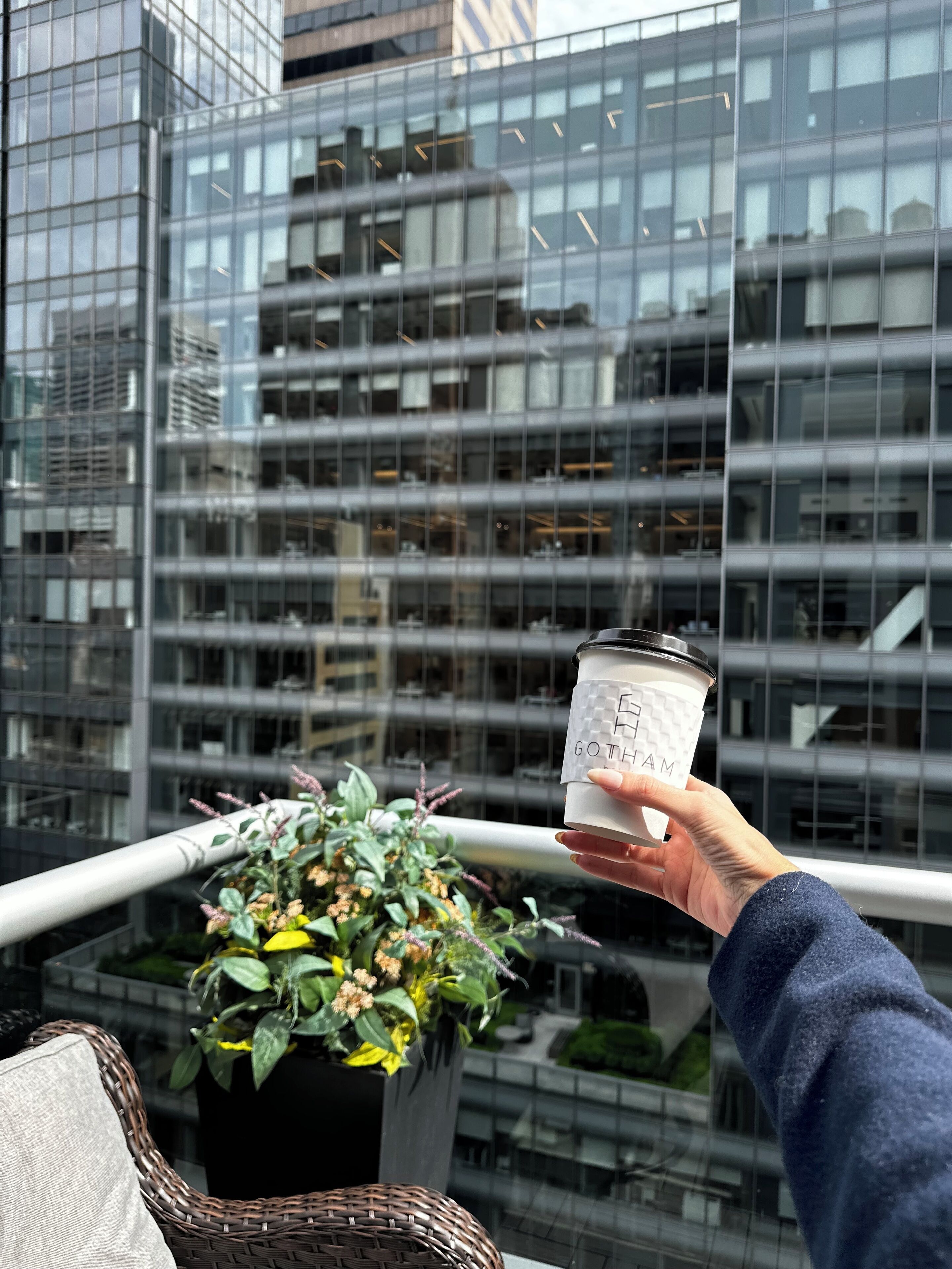 Une main tenant une tasse de café, avec le mot « GOTHAM » inscrit dessus, le tout sur fond de bâtiments modernes en verre.
