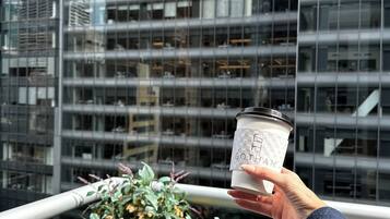 Une main tenant une tasse de café, avec le mot « GOTHAM » inscrit dessus, le tout sur fond de bâtiments modernes en verre.