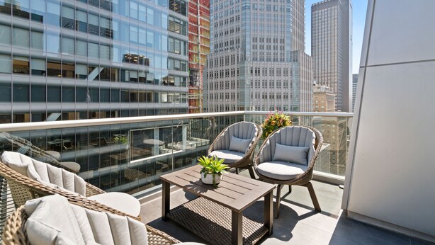 A rooftop terrace with wicker furniture, a small table with a plant, and a glass balustrade overlooking a cityscape with modern buildings. - The Gotham Hotel (New York)