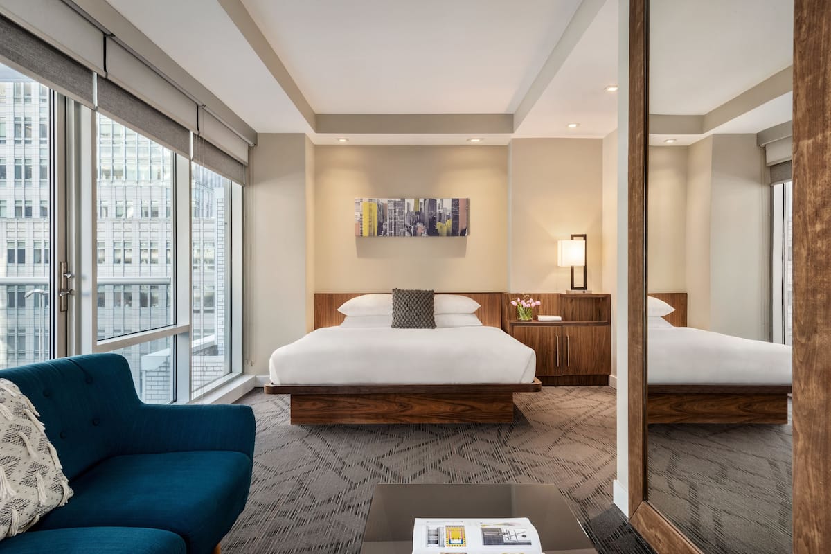 A hotel room with a large bed, a blue sofa, a coffee table, a cityscape view, and a painting on the wall.